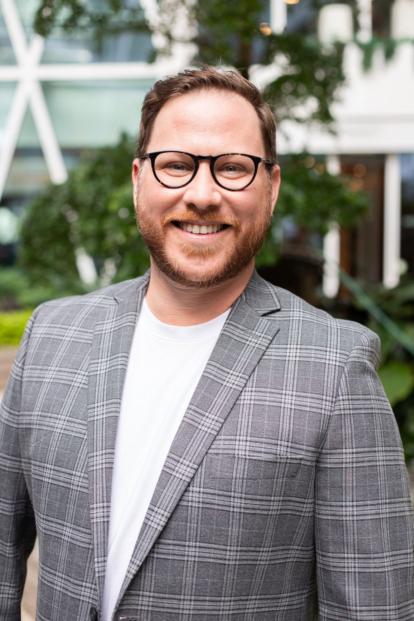 Trevor Paxton, Director of Development. Smiling man wearing round glasses and a gray plaid blazer outdoors, blurred green background behind him.